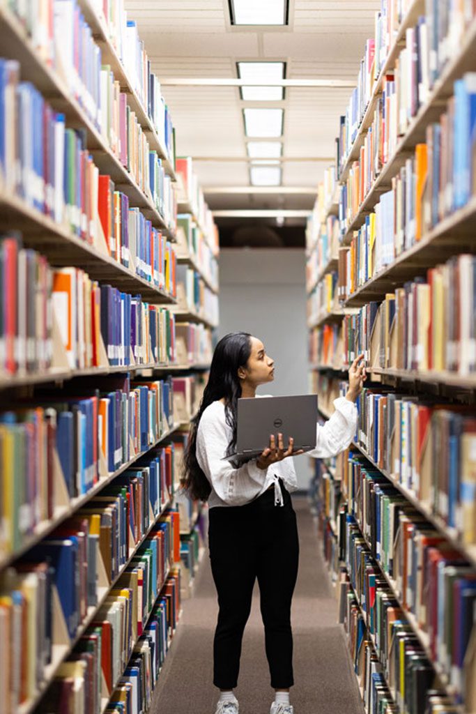 A student holds a laptop while looking at books in the library